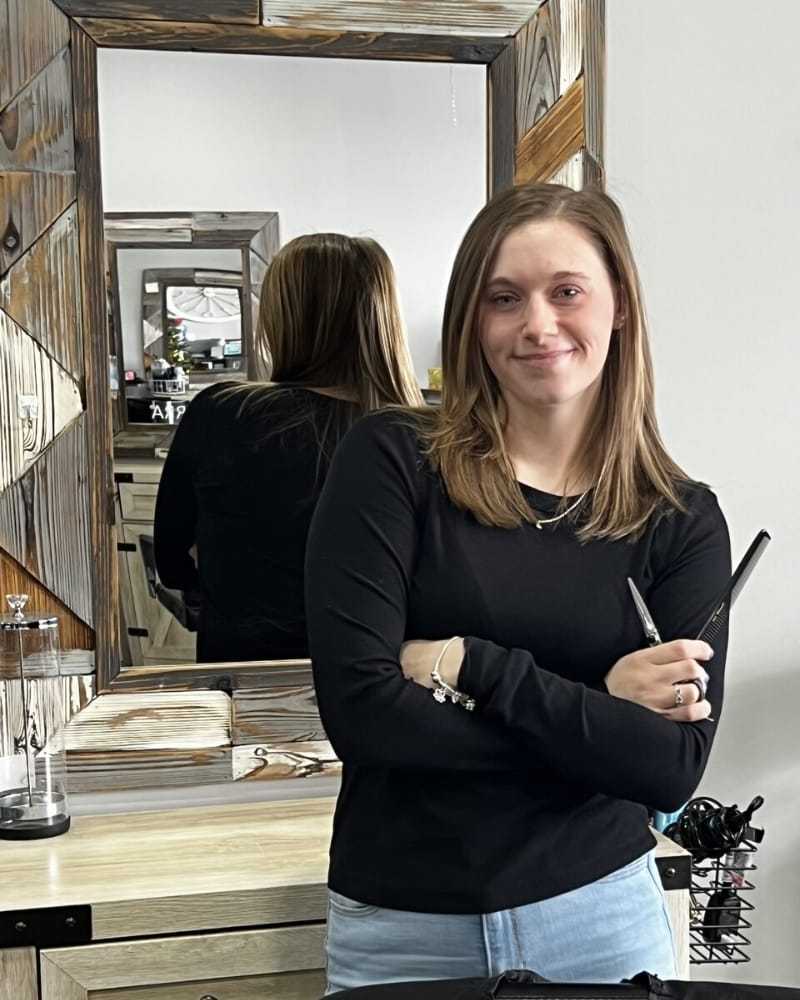 Young woman with light brown hair in salon, holding scissors, stands smiling by a mirror.
